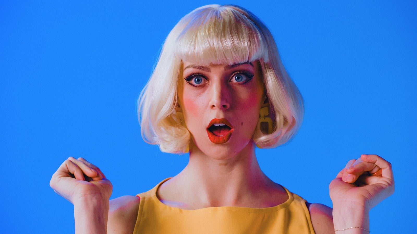 Doll — surprised blonde performer against a blue backdrop
